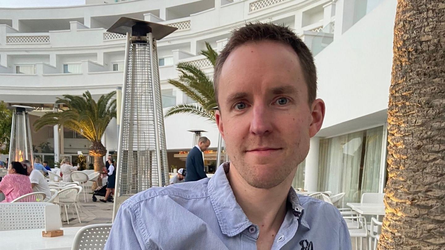 Mark Wherity looks into the camera with a muted expression, he has short brown hair and is wearing a blue collared shirt that is open at the neck. He is sitting down next to a palm tree in an outdoor seating area of a holiday resort