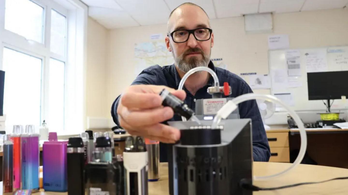 A man with close cropped dark hair and a grey and dark beard dropping liquid from a small black bottle into a table-top device with tubes running out of it. He appears to be in an office and has a number of bottles of vape liquid lined up on a table to his right.