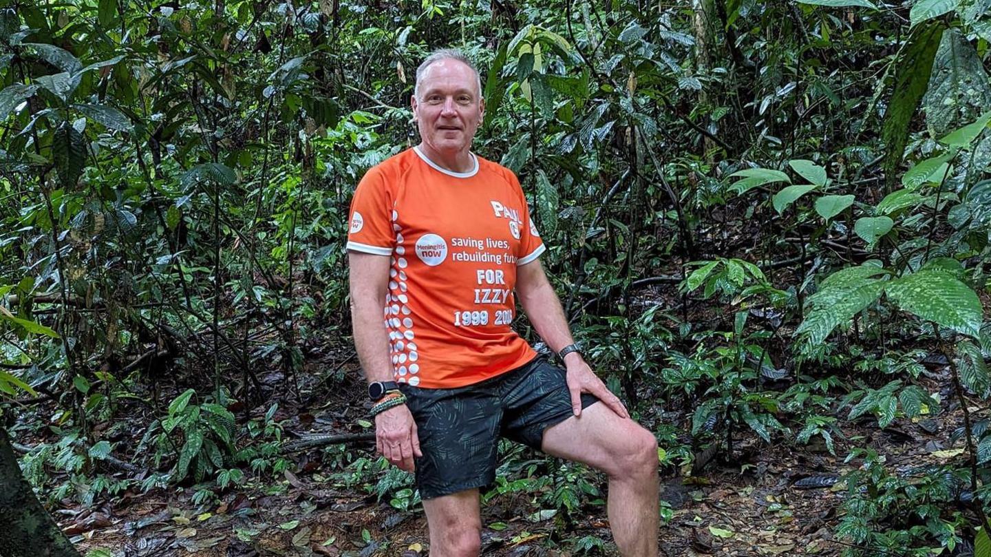 Paul Gentry wearing an orange charity T-shirt and dark shorts looking into the camera while standing in a jungle