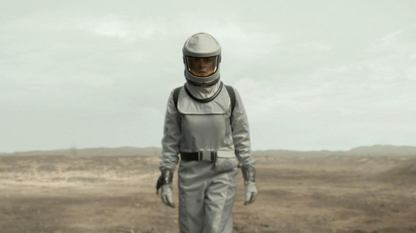 A woman wearing a protective white suit and helmet with a closed visor stands looking directly at the camera with a crater wasteland behind her.