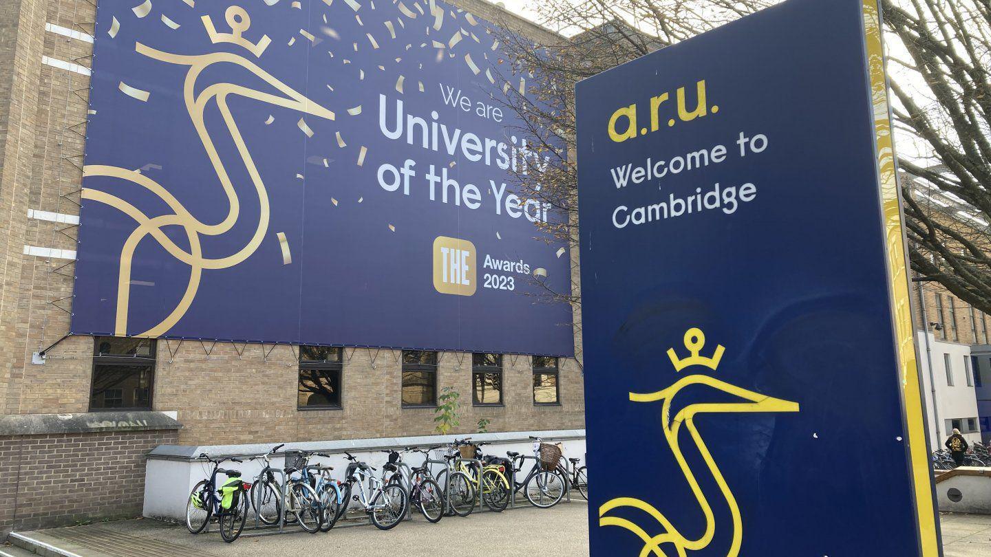 Sign for Anglia Ruskin University. It is blue with a yellow heron insignia on it. Outside the university building are rows of bicycles.