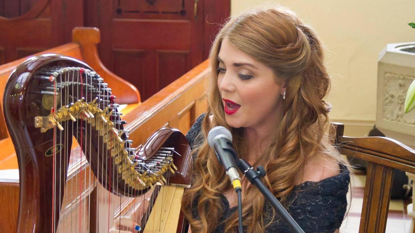 Jenny O'Donnell sits as she plays a harp. The large instrument is to her left, and infront of a church style seat. Jenny's long blonde hair rests on the black formal dress she is wearing. She is also wearing silver earrings