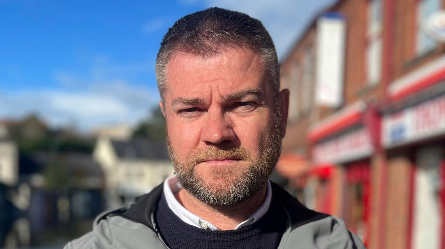 McGrath has short dark hair, a beard and a moustache. He is staring at the camera as he wears a white shirt, black round neck jumper and grey coat.