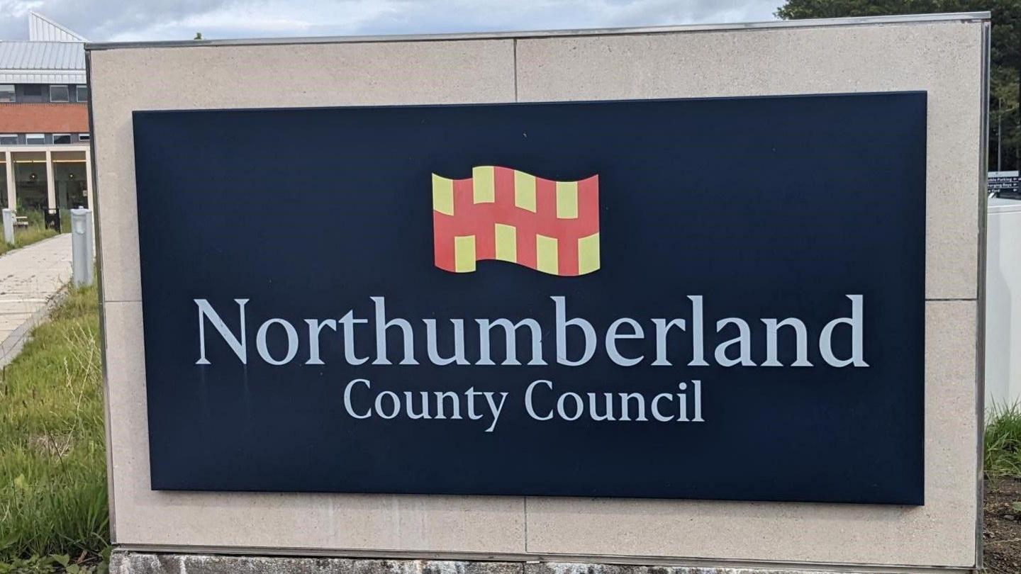 A black sign with white writing which reads 'Northumberland County Council' which sits underneath a red and yellow flag.