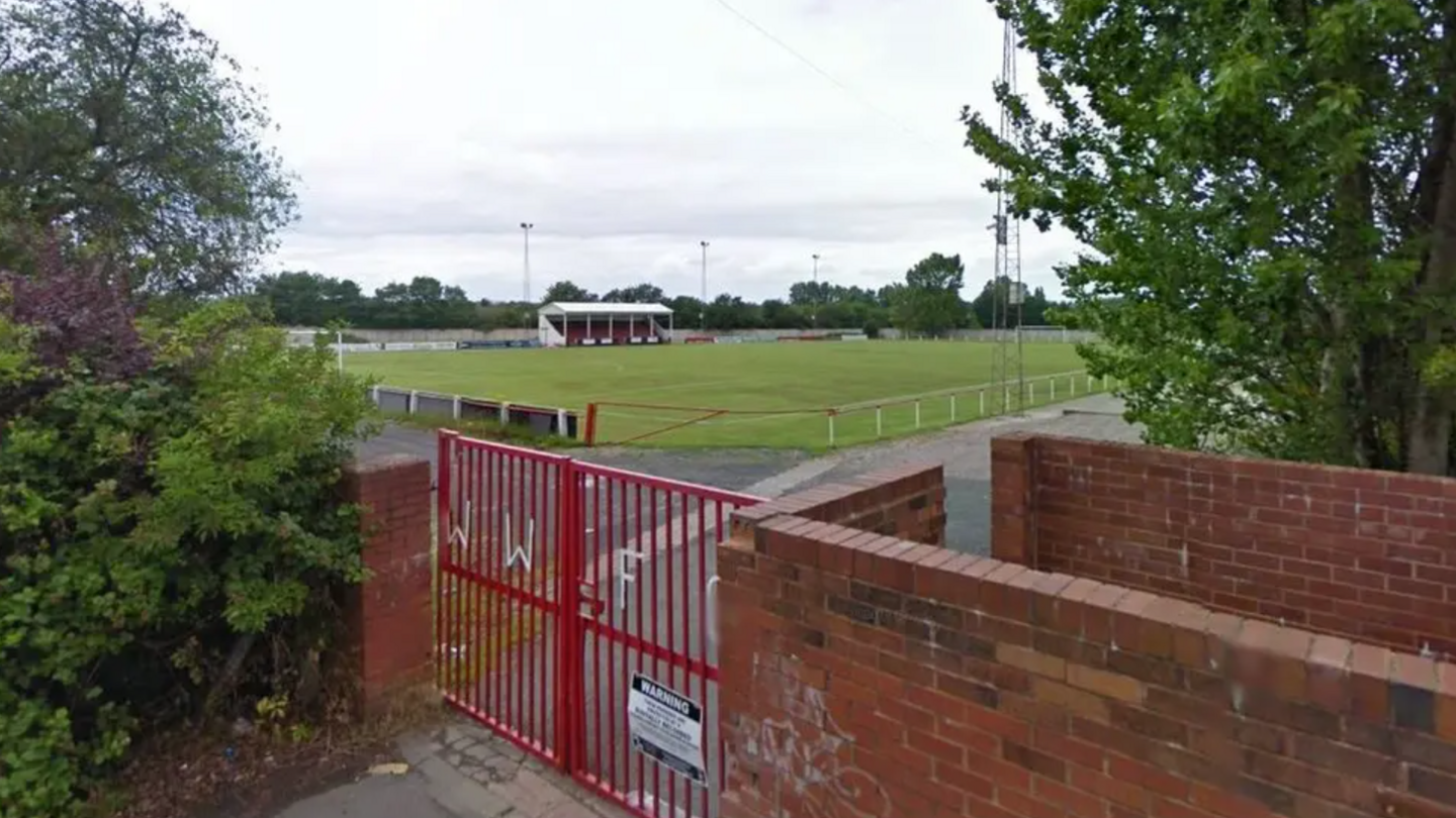 The entrance to Walsall Wood football club. There is WWFC lettering on the fence and a pitch can be seen in the background. 