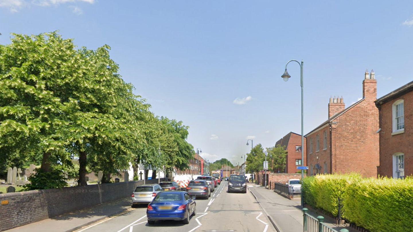 A residential road with houses either side and trees behind a wall on the left. Cars can be seen queuing up on the left.