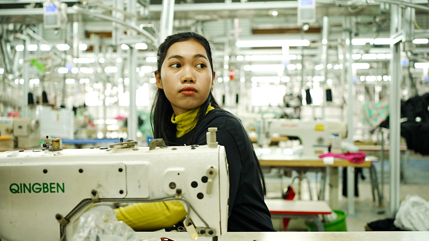 A female worker in a brightly lit factory sits behind a sewing machine. Behind her, more sewing machines can be seen. 