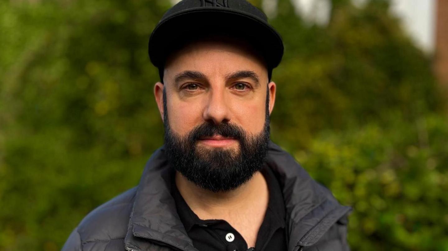 Joe Hall, looking directly at the camera in front of greenery near his house. He has a black cap on. Mr Hall has a black beard and is wearing a navy puffer jacked with a black polo shirt on underneath