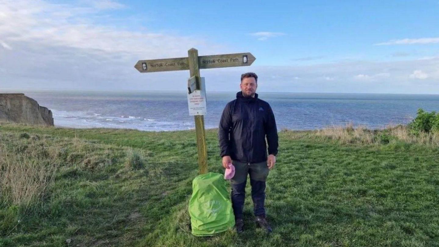 Paul Harris dressed in dark hiking clothes, standing next to a wooden fingerpost. In the background is the sea.