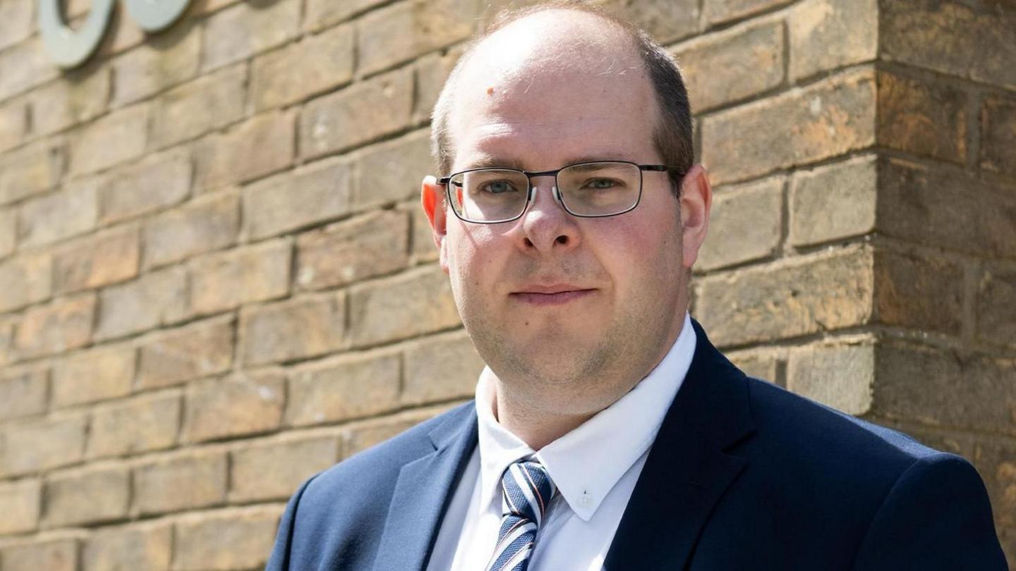 Jonathan Ash-Edwards is wearing a navy suit jacket with a white shirt and a blue, red and white striped tie. He is stood outside against a brick wall and is wearing glasses and looking at the camera smiling.