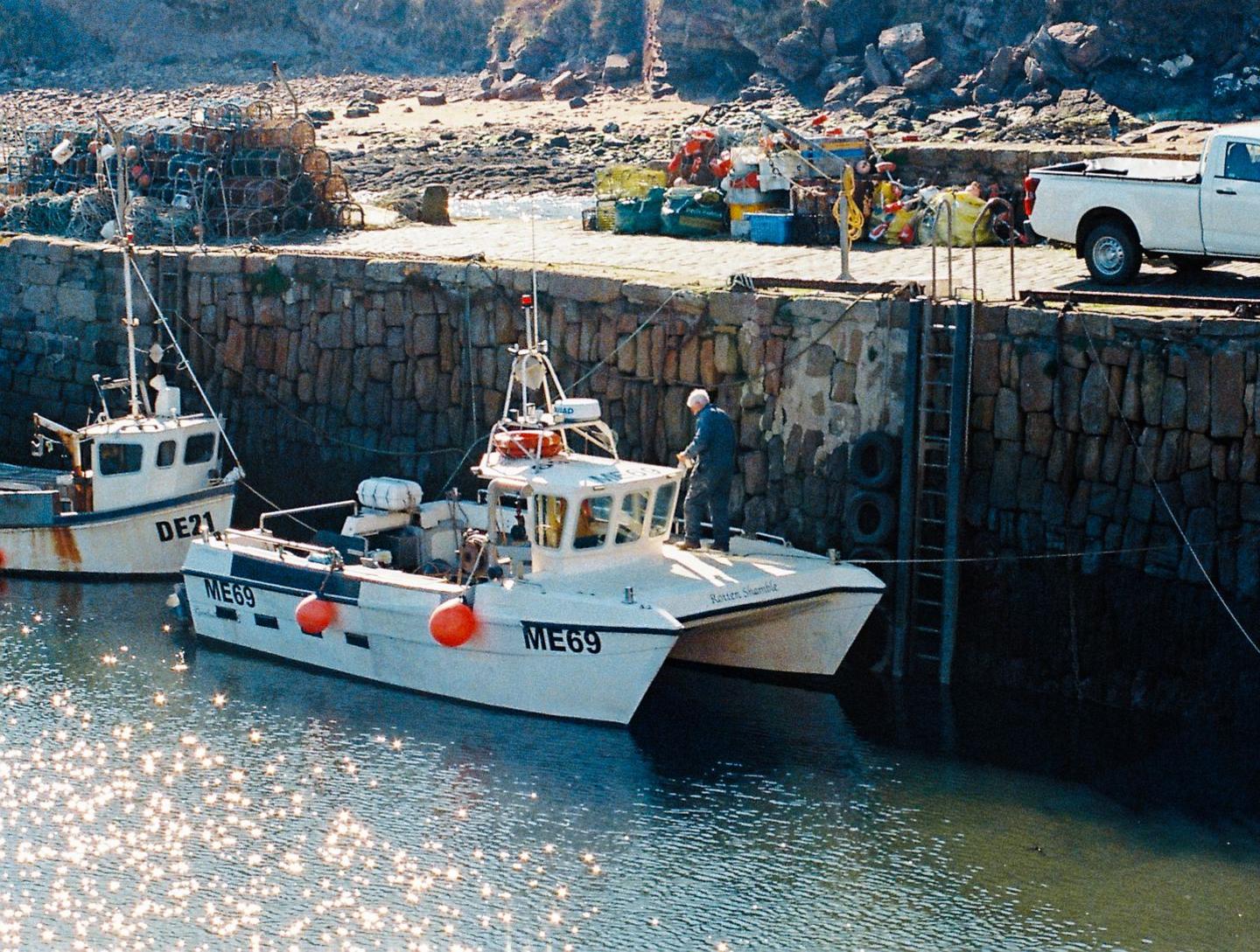A man stands on a small boat by reflective water. Above him, accessible by a nearby ladder, is the walkway of a harbour with nets and lobster boxes.