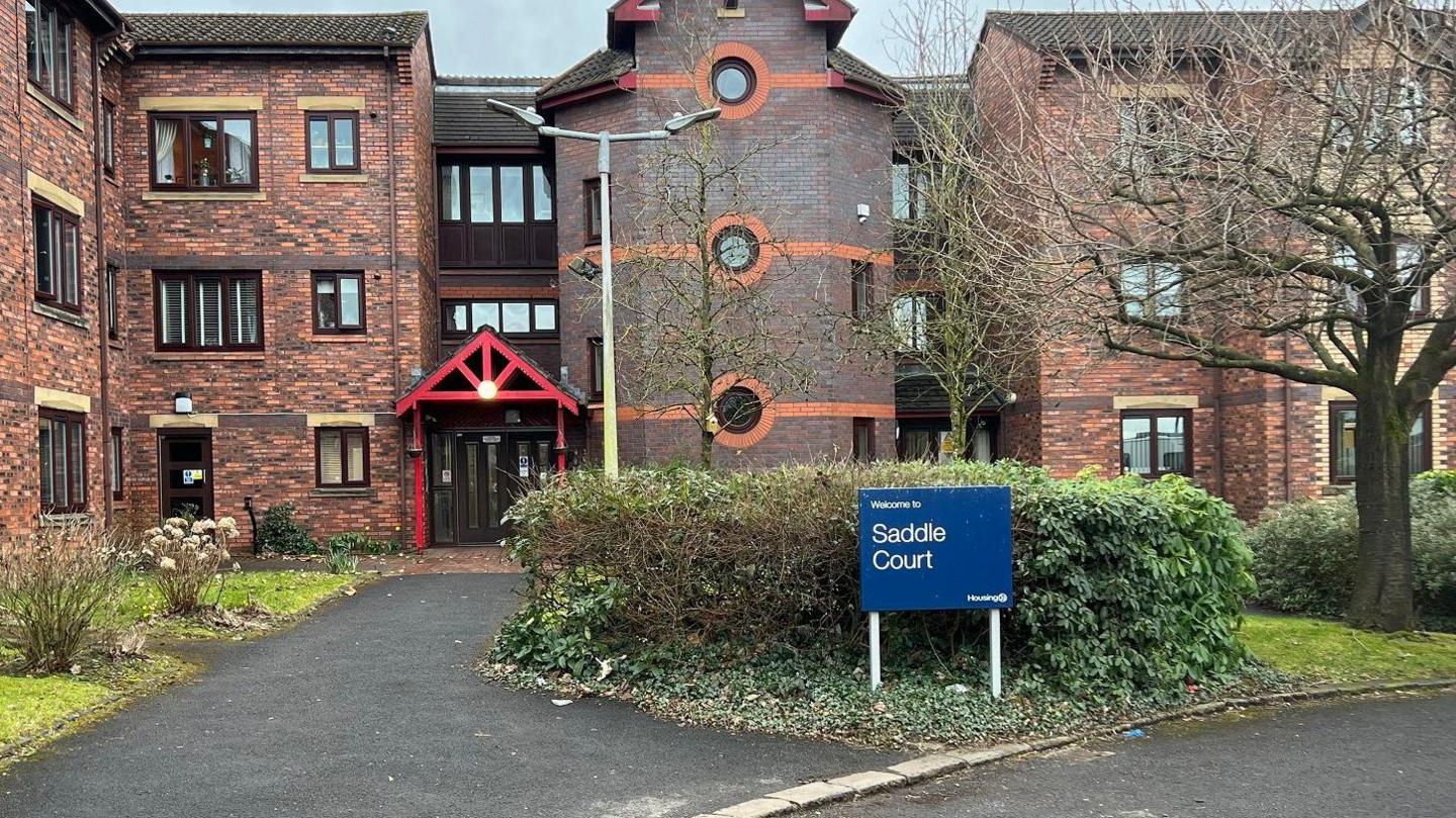 A brick-built block of three-storey flats, with bushes outside and a blue sign that says "Welcome to Saddle Court".