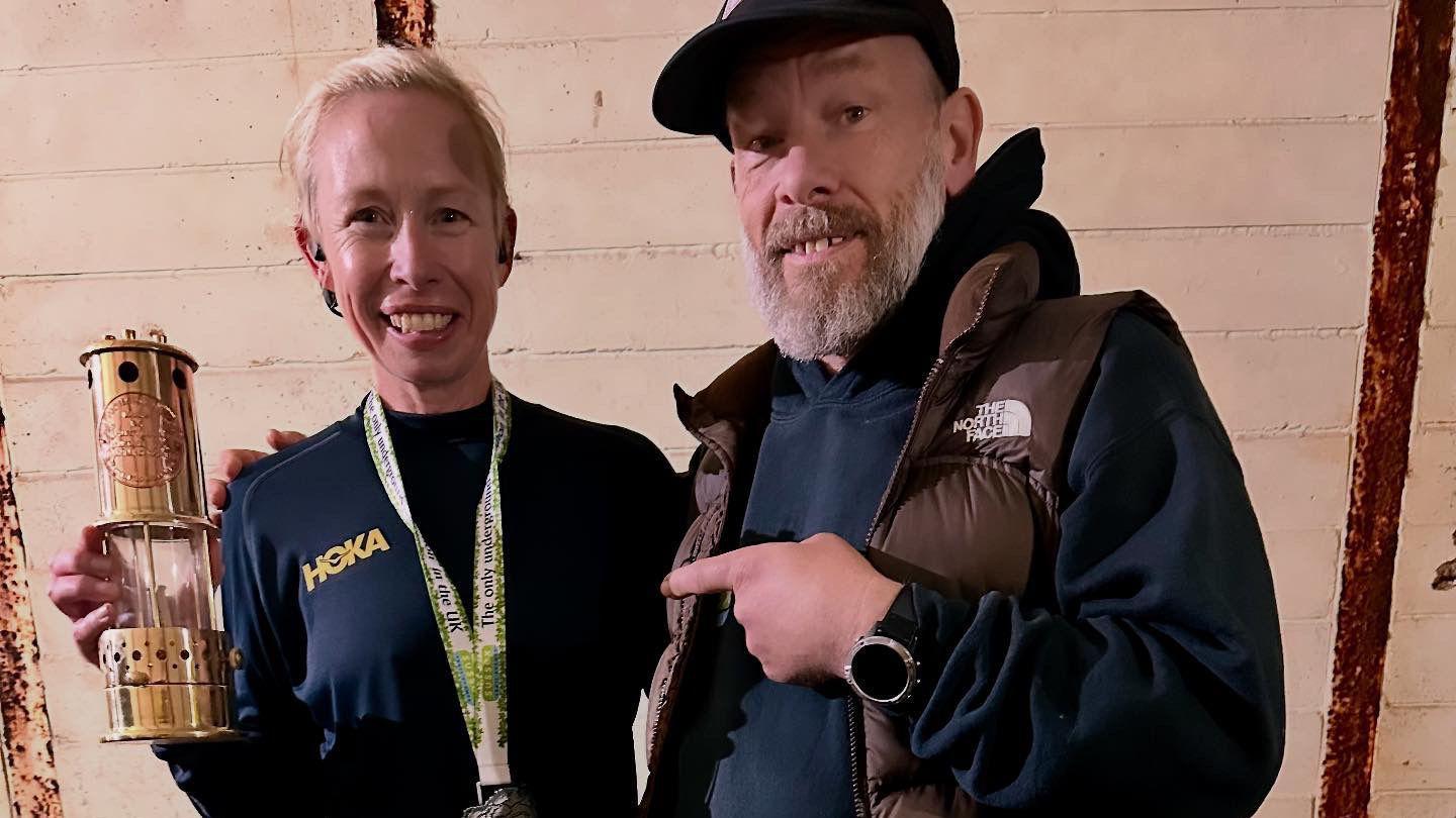 A smiling Laura Watts with Jay McCardle. Laura, who has blonde hair, is holding up her marathon trophy, a miners oil lamp.