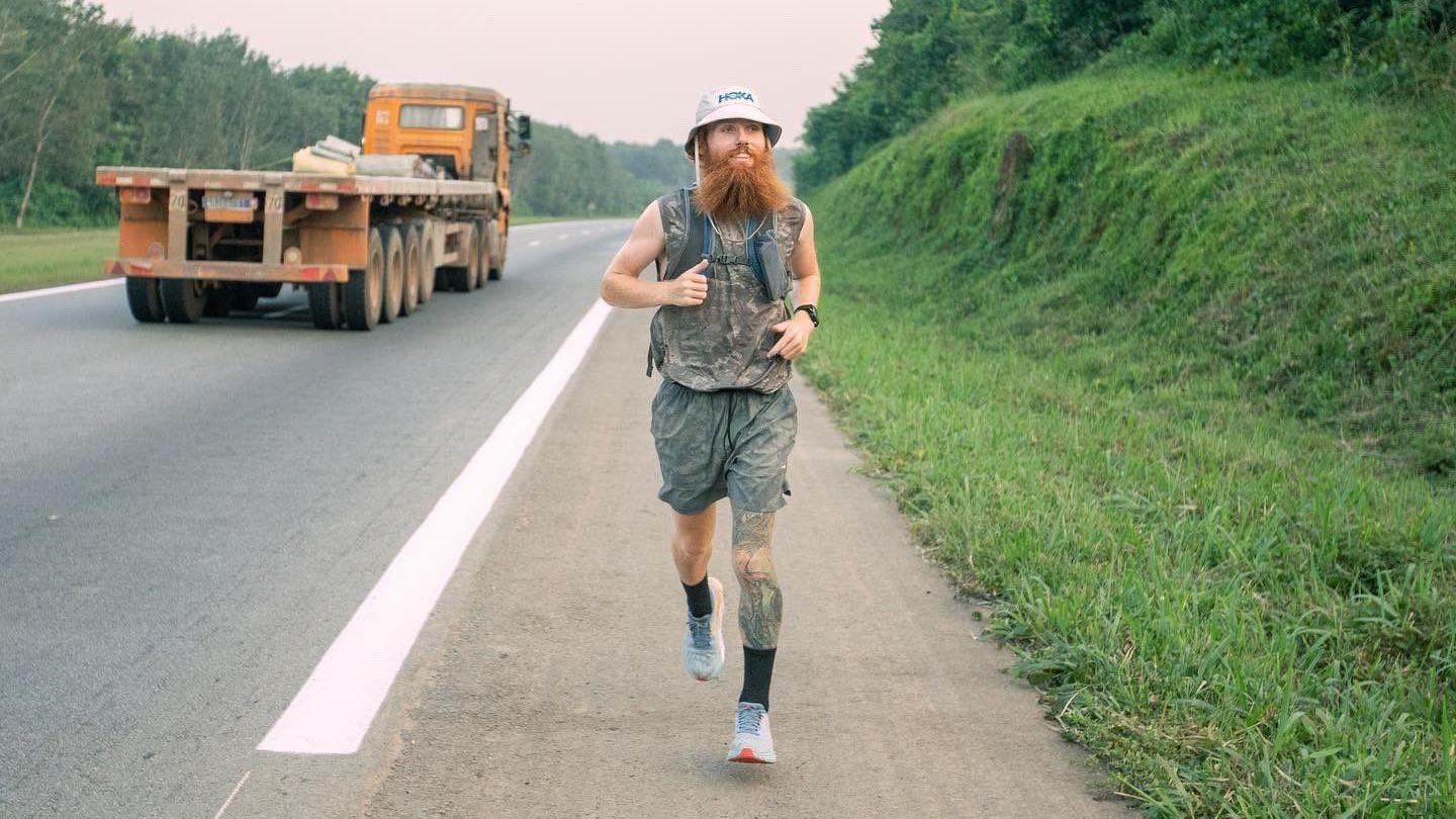 Russ Cook running along the site of a road in Africa. He has a bucket hat on and large ginger beard. He is wearing a sleeveless top and shorts. 