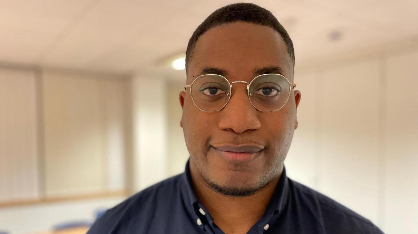 Oba is stood in an office at the BMA offices in Cardiff, behind him are white walls and office desks and chairs. He is wearing a navy shirt and metal-rimmed glasses. It is a head and shoulders shot.