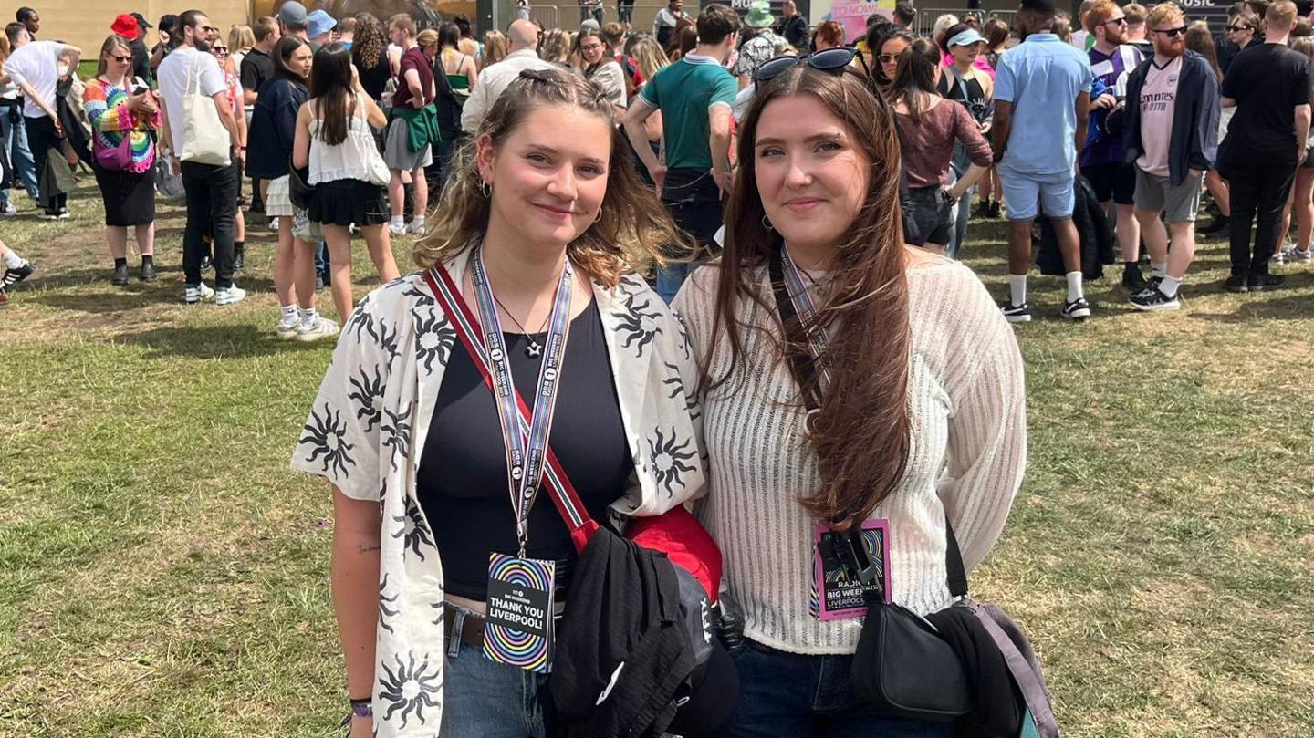 Kate and Scarlett, two friends, standing in a green field. They are smiling. Kate is wearing a black top with a white overshirt with a floral pattern, while Scarlett is wearing a white top.