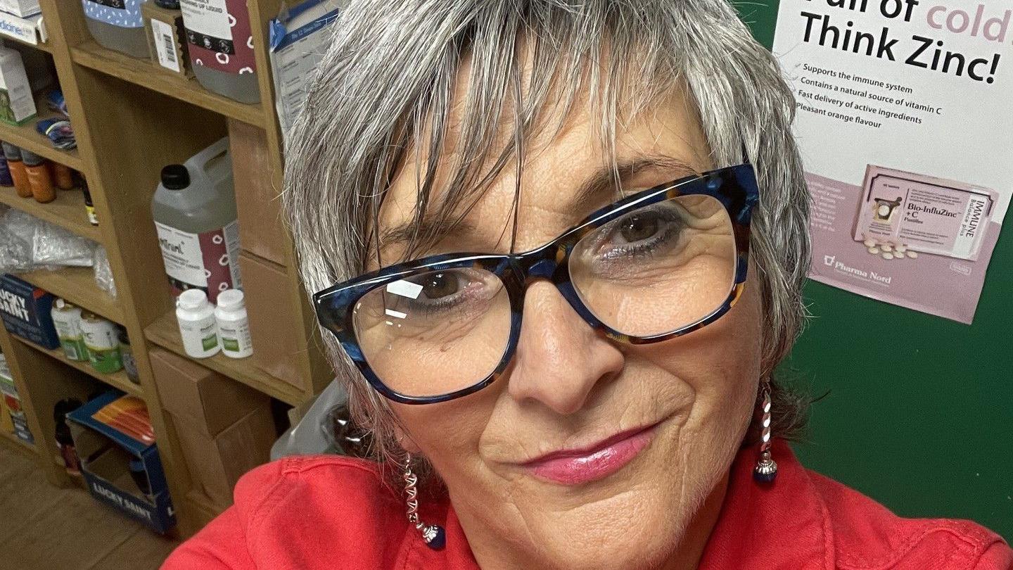 Sue seen with short hair, glasses and wearing a red jacket with earrings