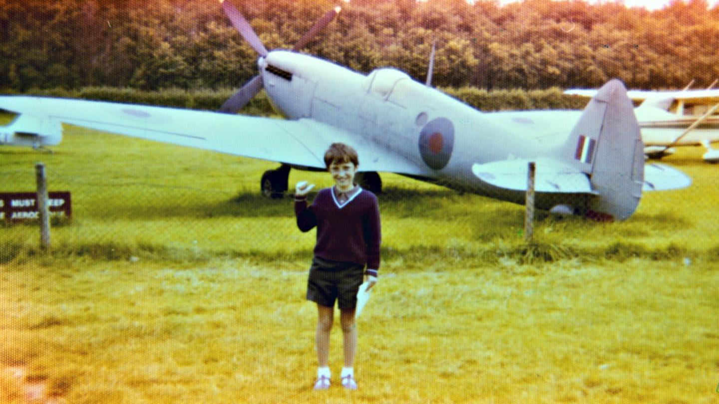 A nine year old boy wearing a deep maroon v-neck jumper and shorts, with white socks and buckle up shoes, is standing in front of an old aircraft. His right thumb is pointing at the aircraft and he is smiling at the camera.