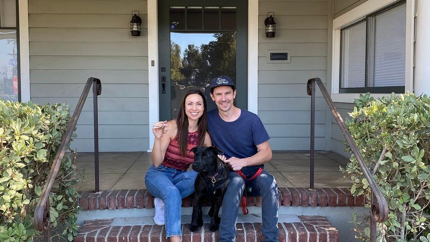 A picture of the couple and a pet dog sitting on the front step of their home before it was destroyed. They are both smiling directly at the camera. The house is painted duck egg blue and there is shrubbery on either side of the steps.