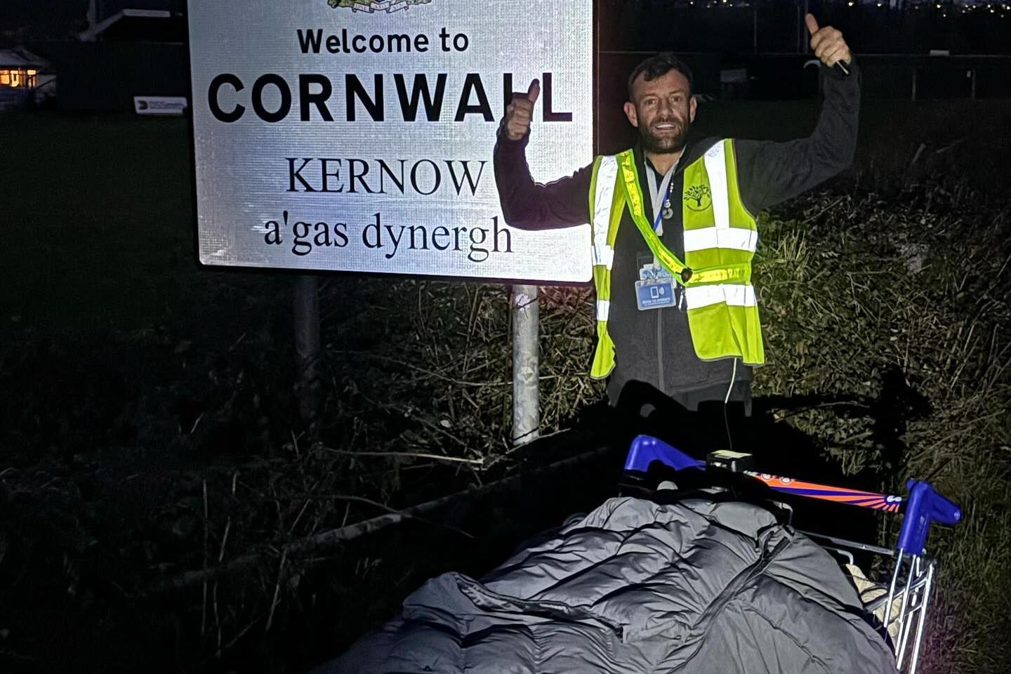 Perry Pel Scopes stands in front of the Welcome to Cornwall sign. It is dark and he is wearing a high vis vest over a jumper. His shopping trolley is in front of him. 