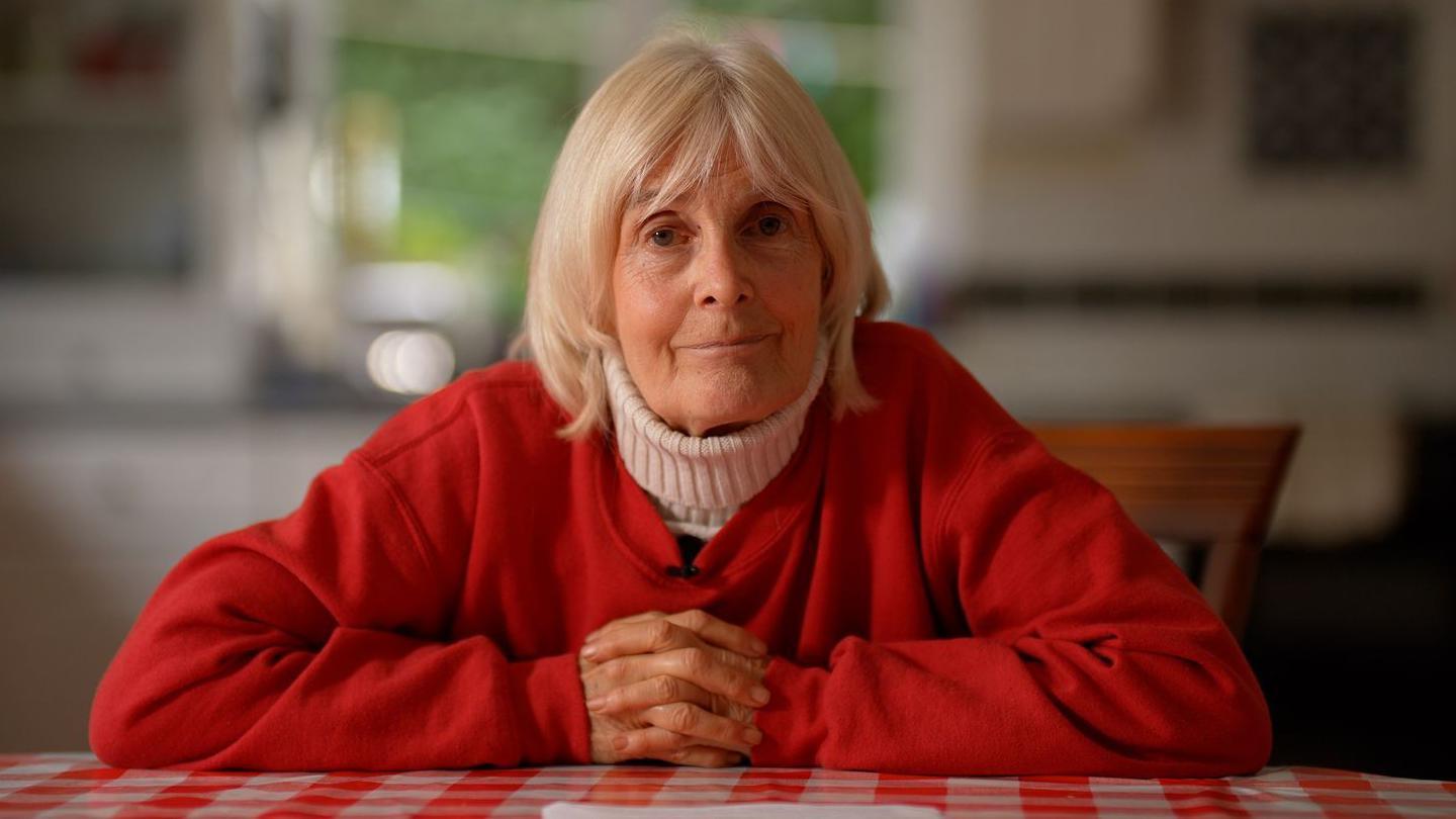 Maureen Treadwell in a red jacket and white cardigan sat arms folded at her kitchen table