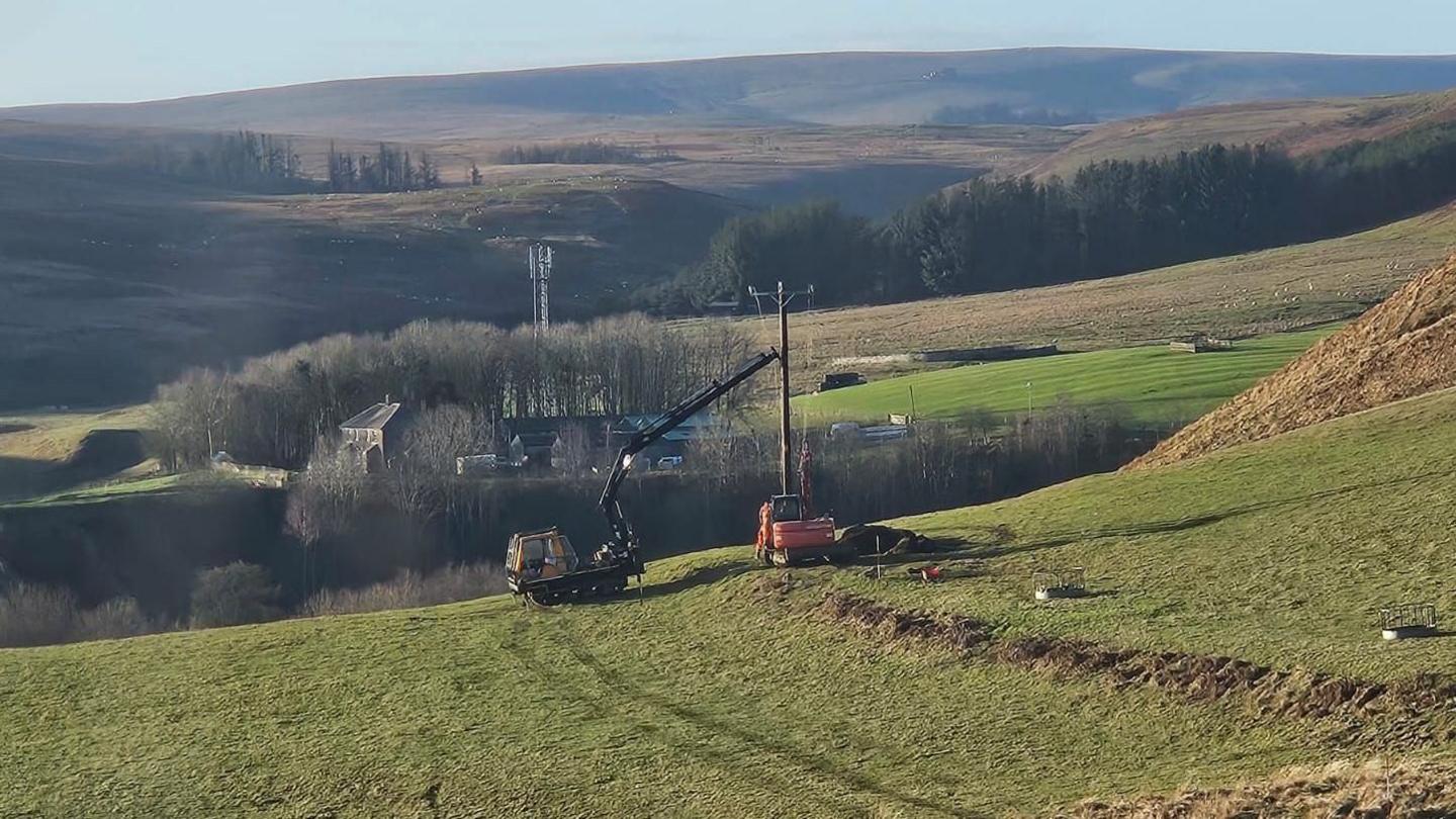 A pole is being put up by a truck with a long extendible arm which is holding it in place. In the distance there is a farm and a telecommunications mast, while desolate hills poke up in the distance.
