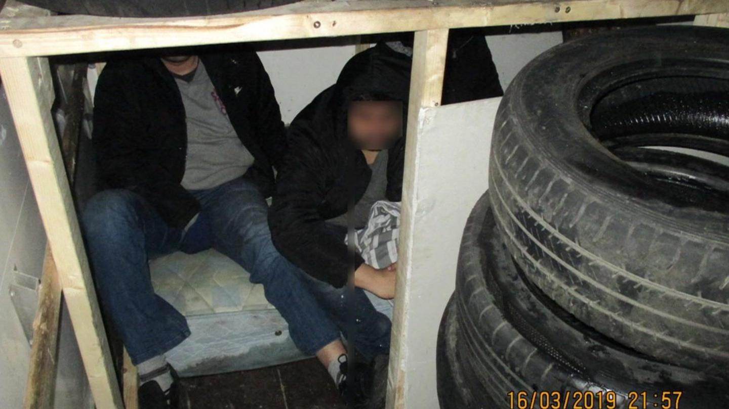 Two men are sitting inside a confined compartment constructed with wooden panels in the back of a van. The men are partially obscured, wearing dark clothing, and one has a blurred face. The compartment is surrounded by stacked car tyres, and a mattress is visible under the men.