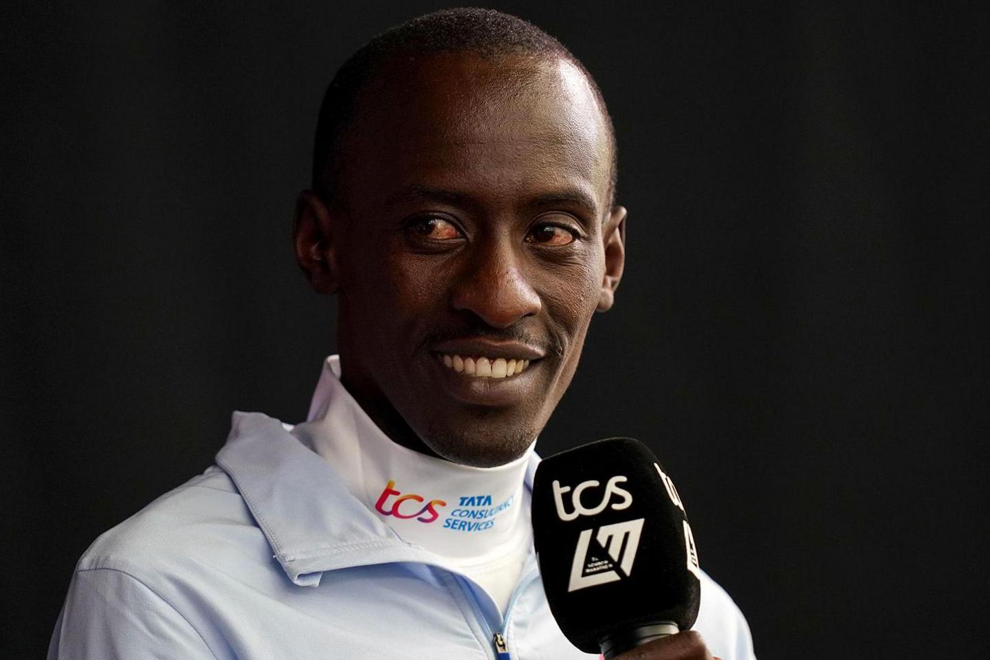 Kelvin Kiptum is photographed against a black background - someone is holding a microphone up to him but he is looking away and smiling at something beyond the camera. He wears a lightweight blue zip-up jacket over a high-necked white top with sponsorship branding on it