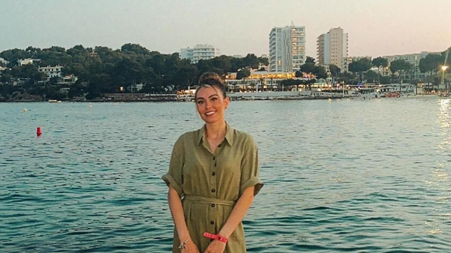 Cheryl Bennett, who has her blonde hair in a bun, is wearing a green dress as she smiles at the camera. She is stood in front of the sea in late afternoon or early evening.