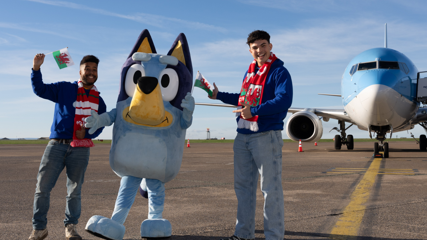 Bluey standing beside two men. They are wearing jeans, blue hoodies and Wales scarves and are holding Welsh flags. All three stand in front of the aeroplane.