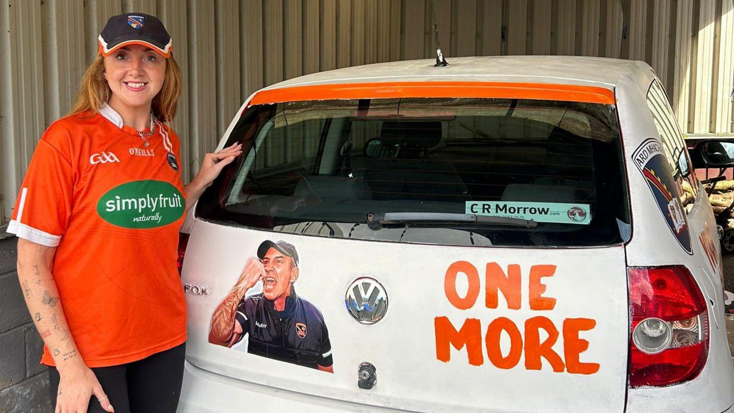 Gillian Kelly, wearing orange Armagh jersey, stands beside her now-orange and white coloured car which has a photo of the manager and the phrase 'one more'