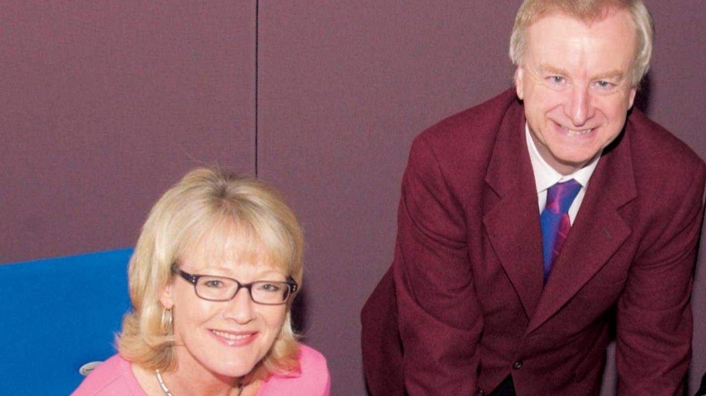 Wendy Austin, who has blonde hair and is wearing glasses is sitting while Seamus McKee, wearing a burgundy suit, white shirt and blue tie, is standing beside her.