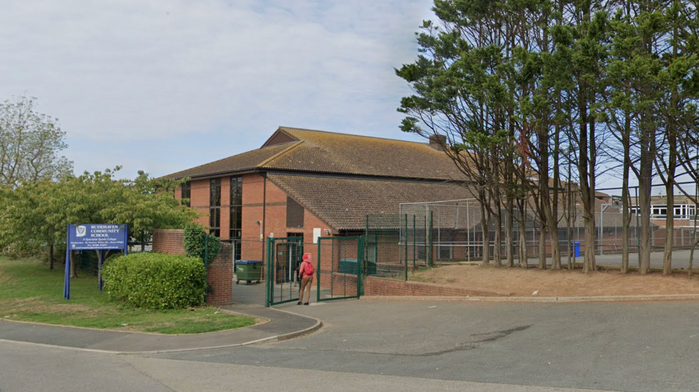 A google maps screengrab of Budehaven Community School, which is a large brown building surrounded by trees and gates, with a blue sign outside and a main road in front.