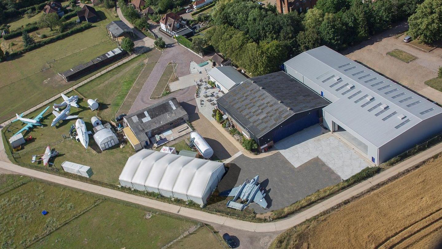 An ariel view of the museum, which includes a sturdy white marquee along with two much larger permanent hangars, one light grey and one dark gery. A number of aircraft are on display on a grassy section. The site is surrounded by fields and some housing.