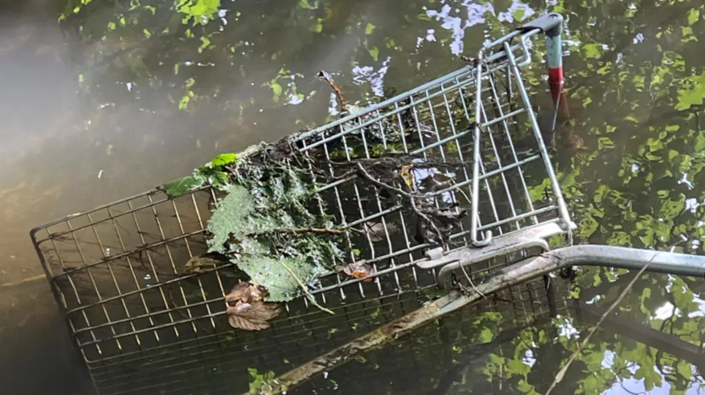 A disused, rusty trolley dumped in the Bottesford Beck. Leaves and algae are intertwined with the metal. Reflections of trees can be seen through the murky brown water.