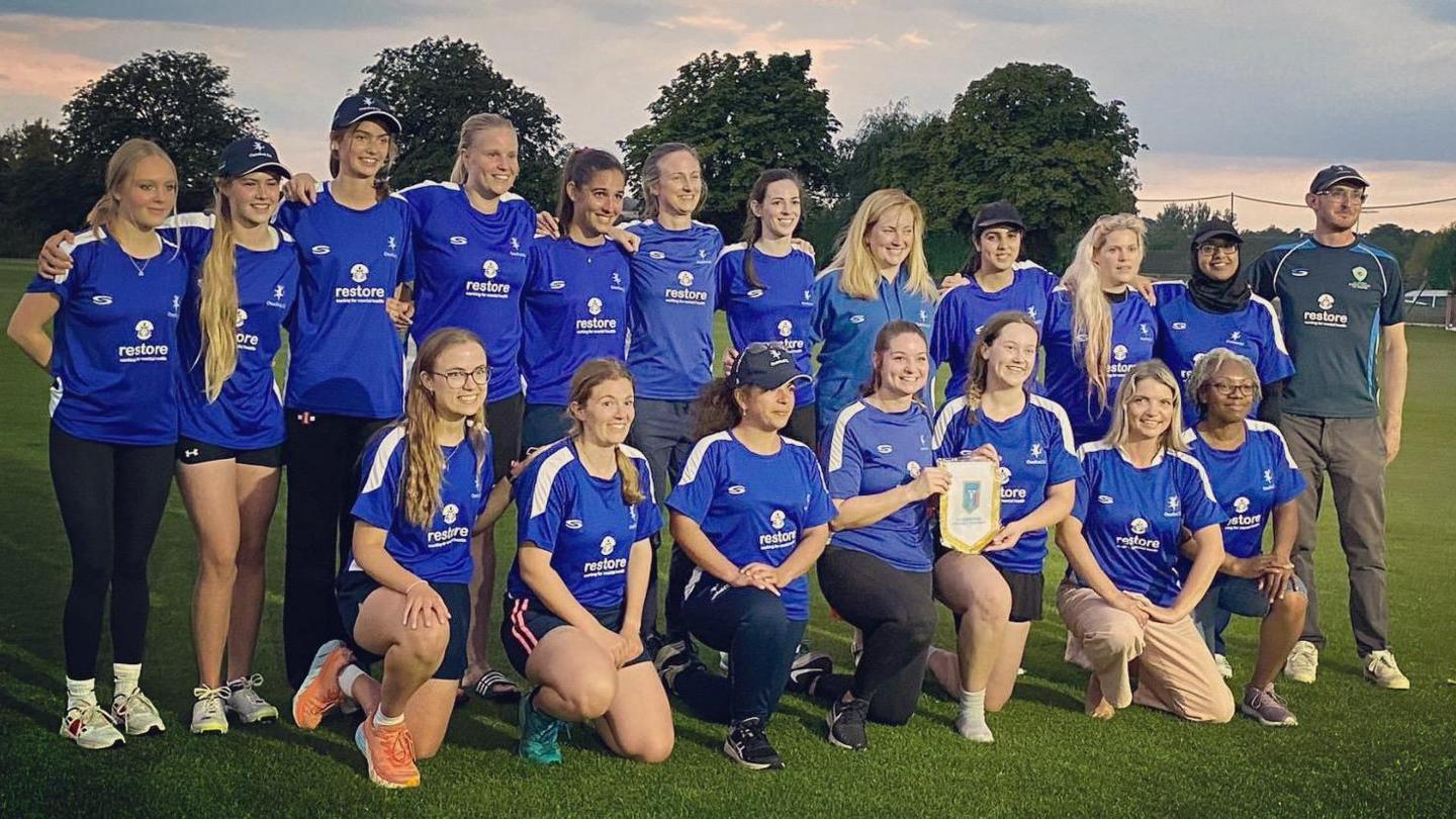 Oxenford CC women players in blue smiling for the camera. The women on the first row are kneeling, while the ones on the second row are standing. Two of the women in the middle bottom row are holding a small flag. There is one man at the end of the second row. It's a cloudy day.