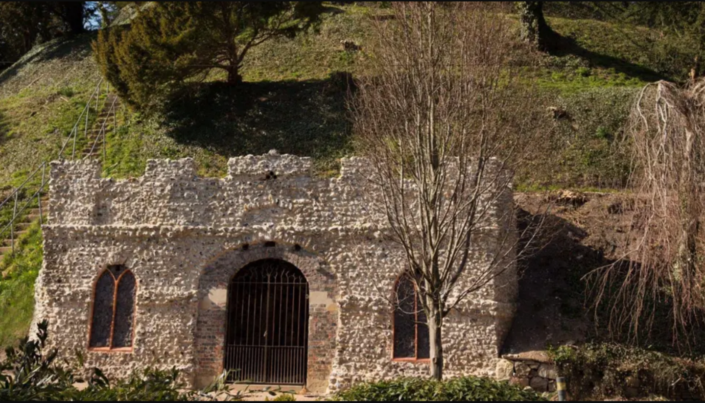 A small, castle-like structure made of small stones and brick with arched church-like windows and an arched gated entrance in the side of the mound.