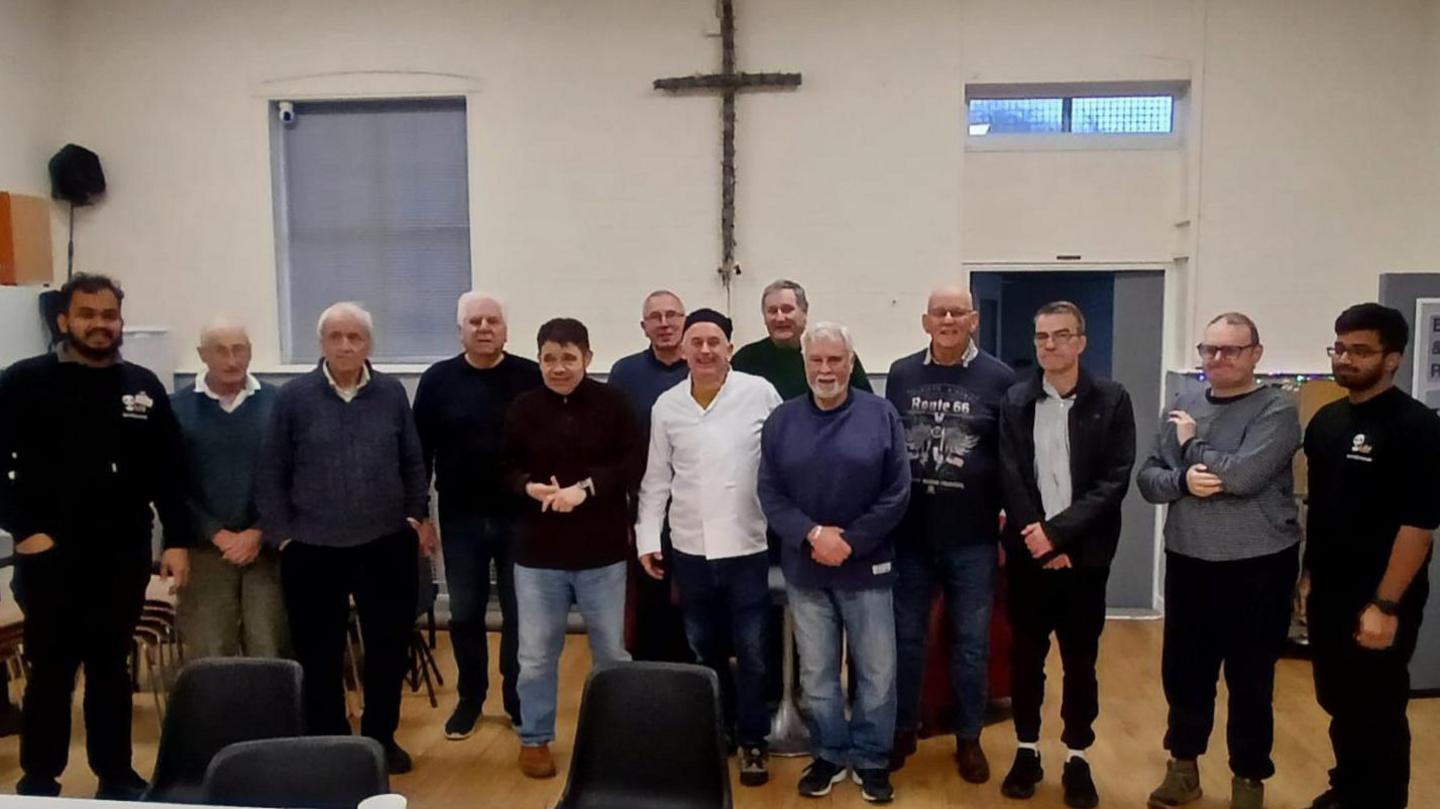 A group of men stand beside one another smiling at the camera. Chairs are pictured in front of them.