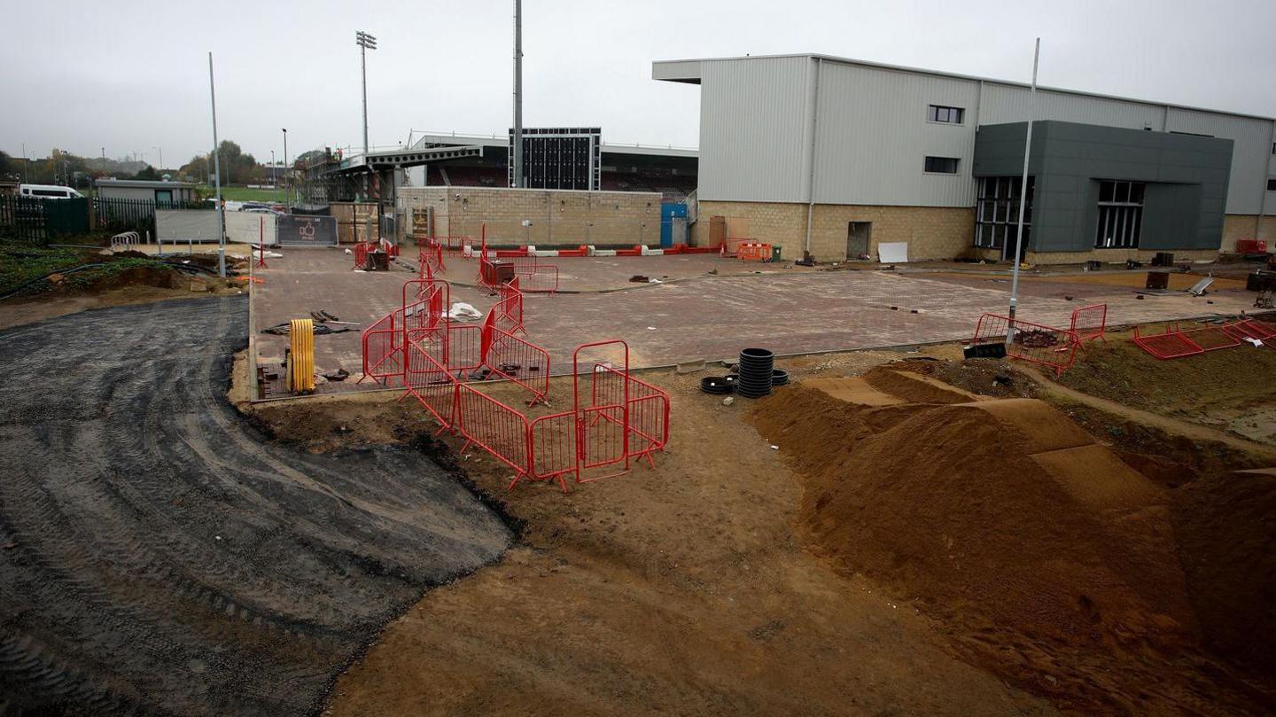 Behind the East Stand at Northampton Town, construction is on going. Big piles of dirt and the start of a tarmac road can be seen. The rest of the stadium is shown in the background. 