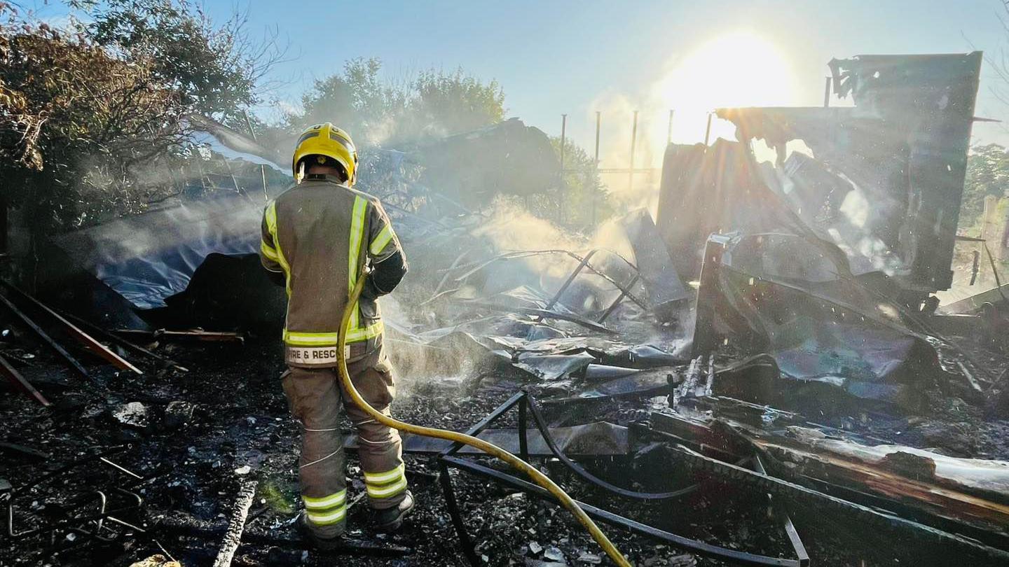 A firefighter hoses down a mangled pile of charred material against a low afternoon sun.