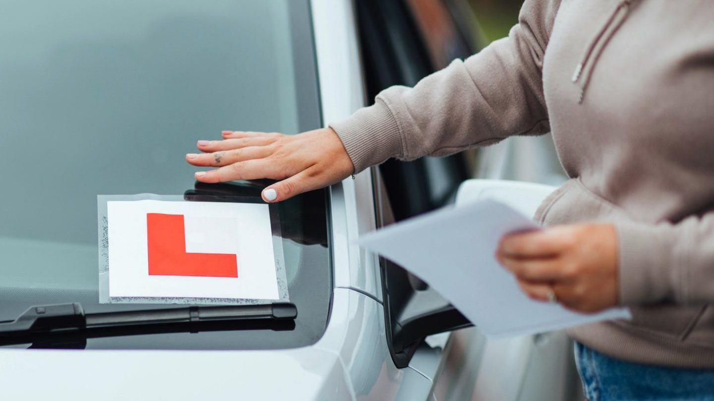 A woman with blonde hair securing a red L plate sticker to the windscreen of her car. She is wearing blue jeans and a faded mauve hoodie.