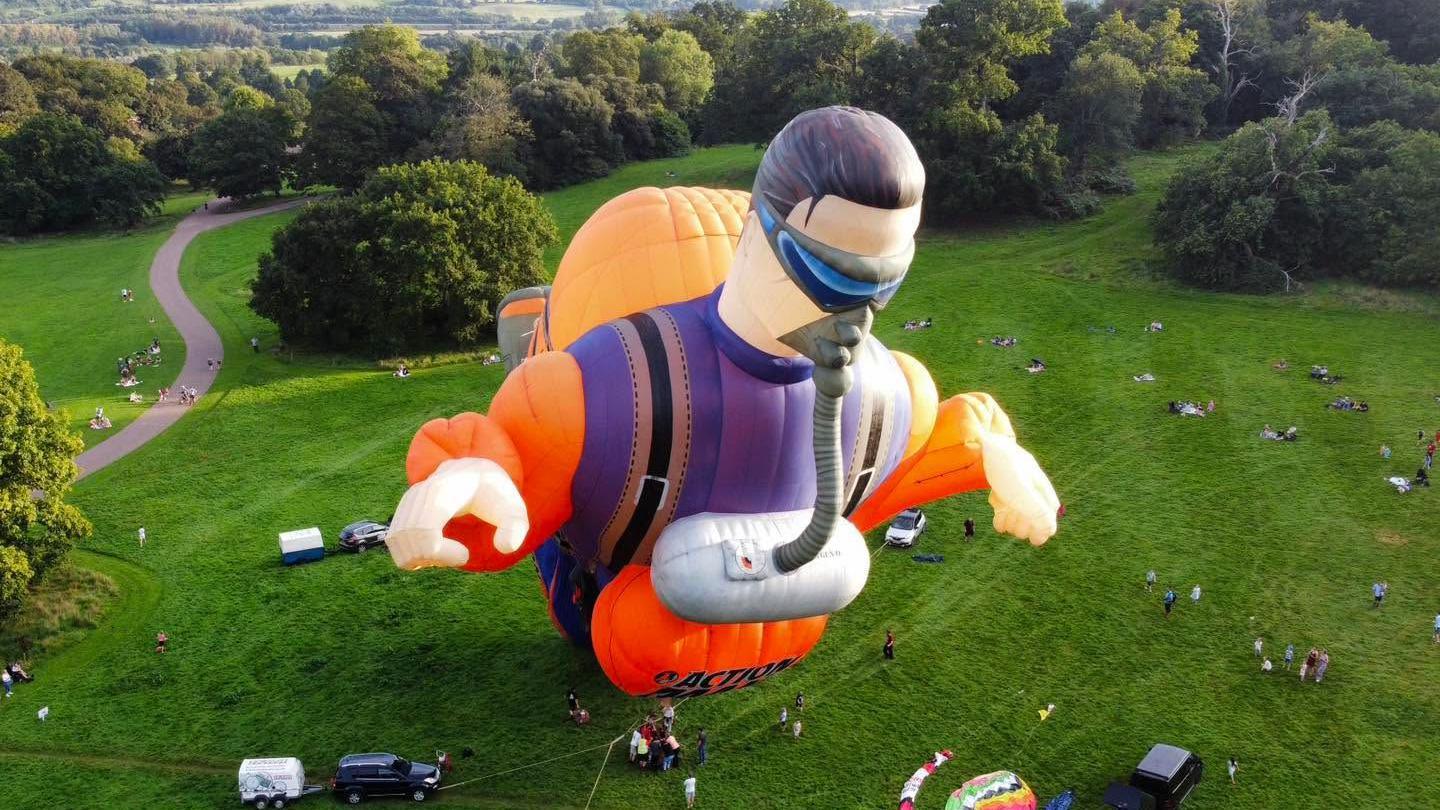 A giant hot air balloon shaped like Action Man tethered to the ground of a large field.