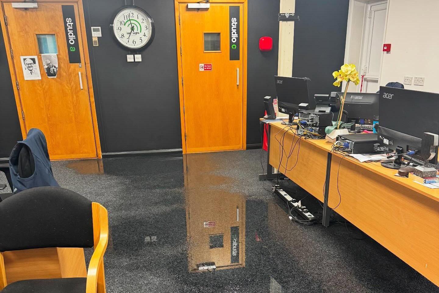 A flooded office. The black floor is covered in water. To the right are two long desks with computers and monitors on them. There are two wooden doors at the back of the room marked Studio A and Studio C.