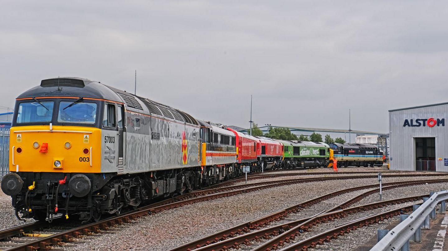 A line of colourful locomotives rests on curved tracks outside an industrial building marked "Alstom"