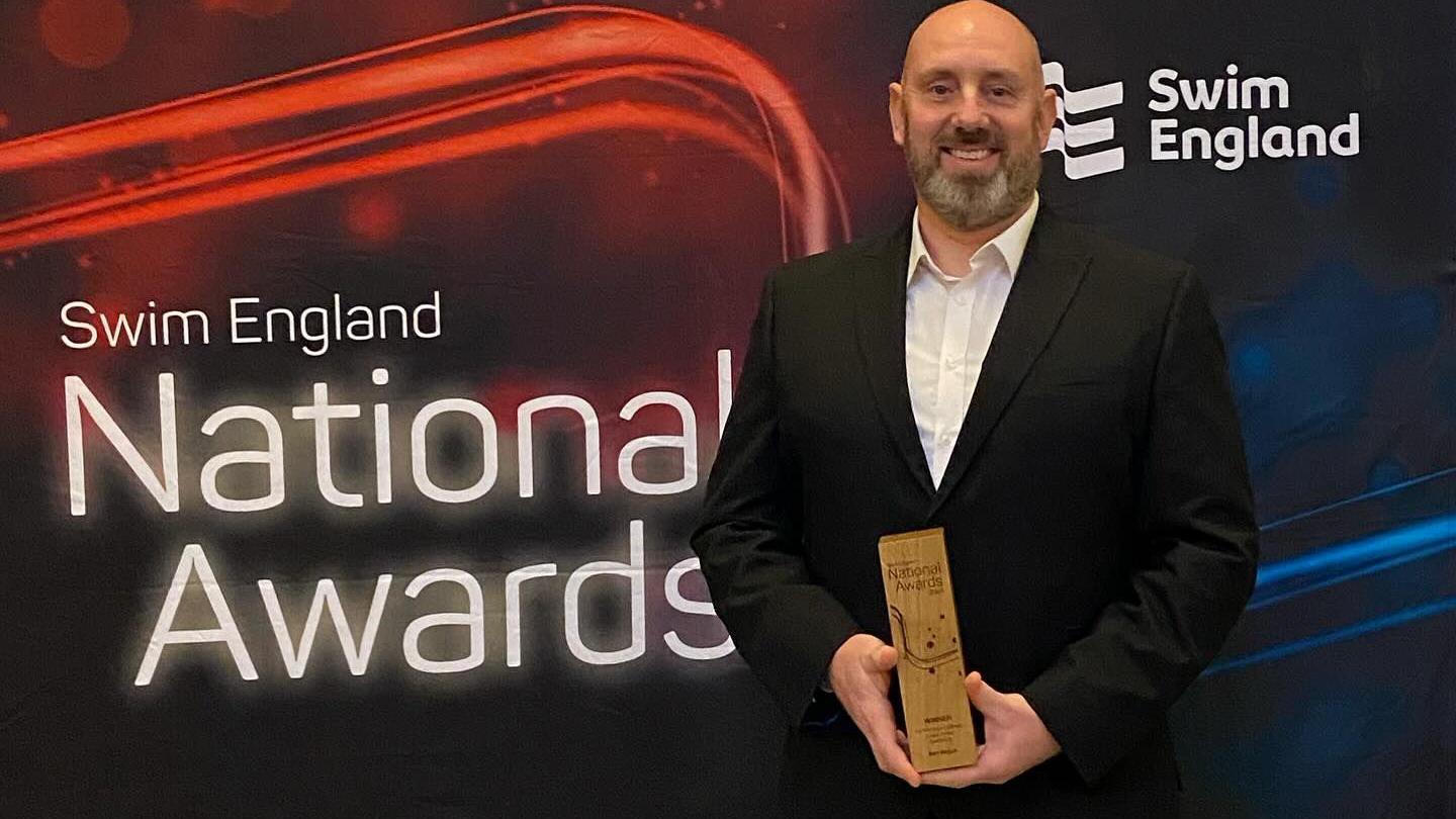 Ben wearing a black suit holding a wooden award standing in front a red and black Swim England banner 