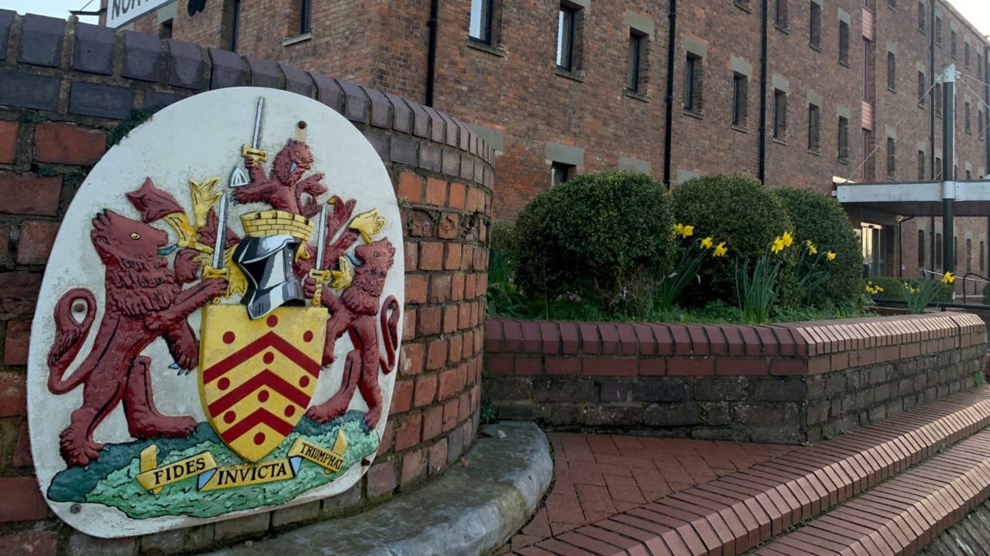 The emblem of Gloucestershire County Council is a coat of arms with chevrons, fleeces, horseshoes, and a crest. The motto is "Prorsum Semper", which translates to "Ever forward". 