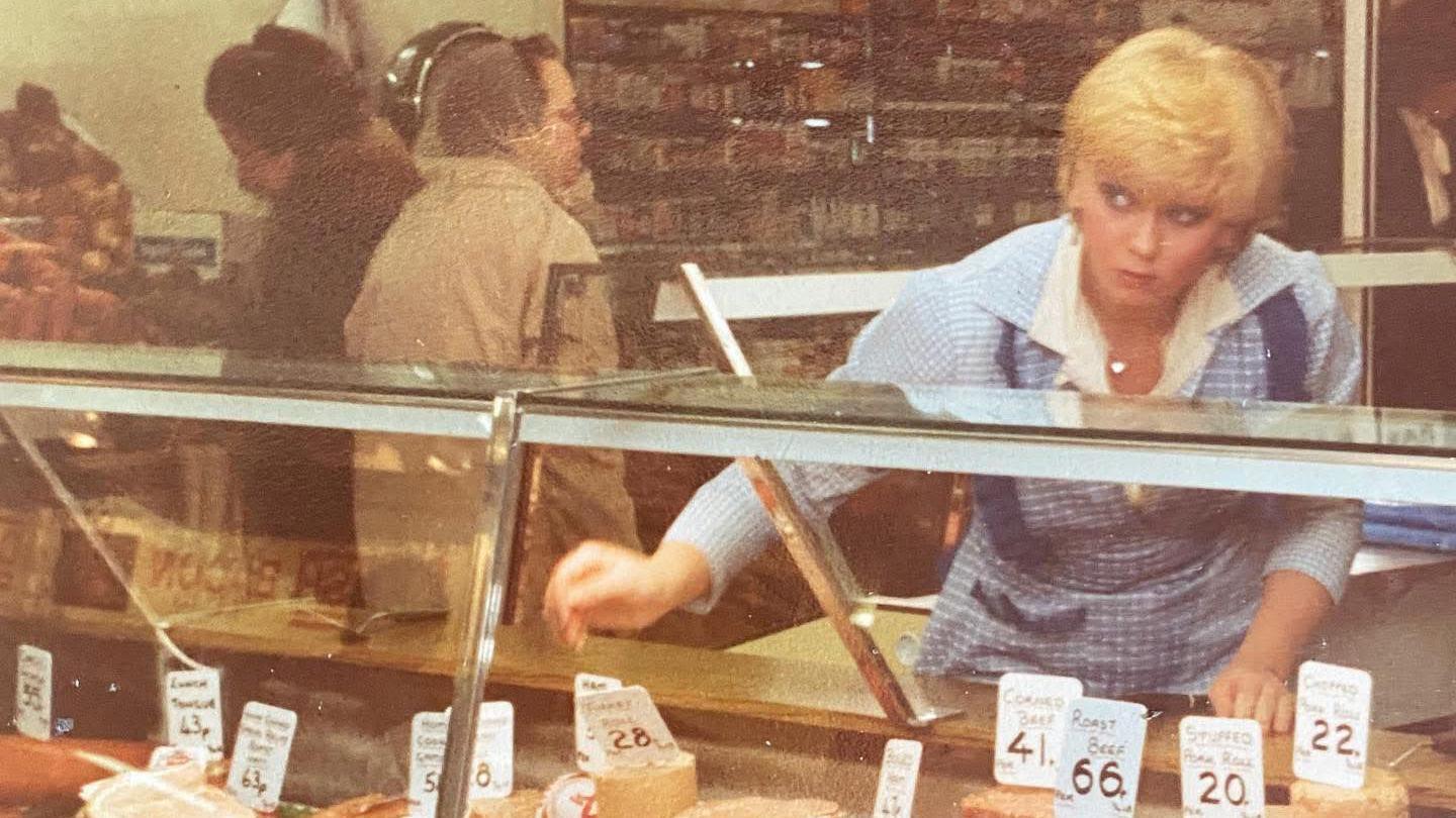 Debbie Hughes leans over a counter of cheese while wearing a blue apron in a dated photo from the market.
