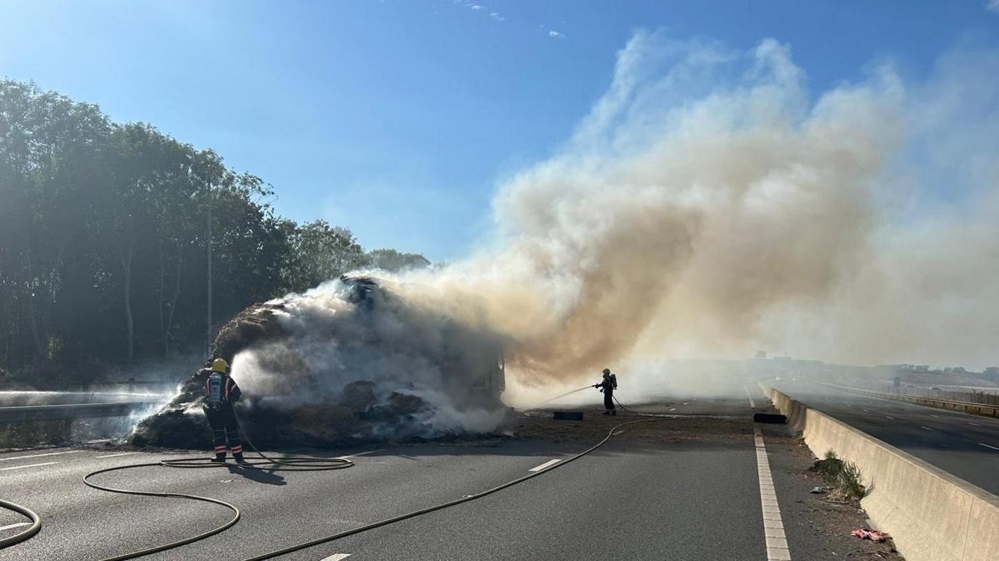 An empty dual carriageway with a large object engulfed in fire. Several firefighters stand nearby pointing hoses towards the fire. The sky is blue and there are trees on the left. The right carriageway is empty.