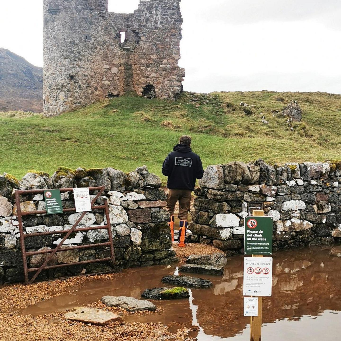 A man stands in an entrance in the wall to the castle and the large puddle with the stepping stones in it. There are warning signs in the foreground and the ruined castle looms large in the distance.