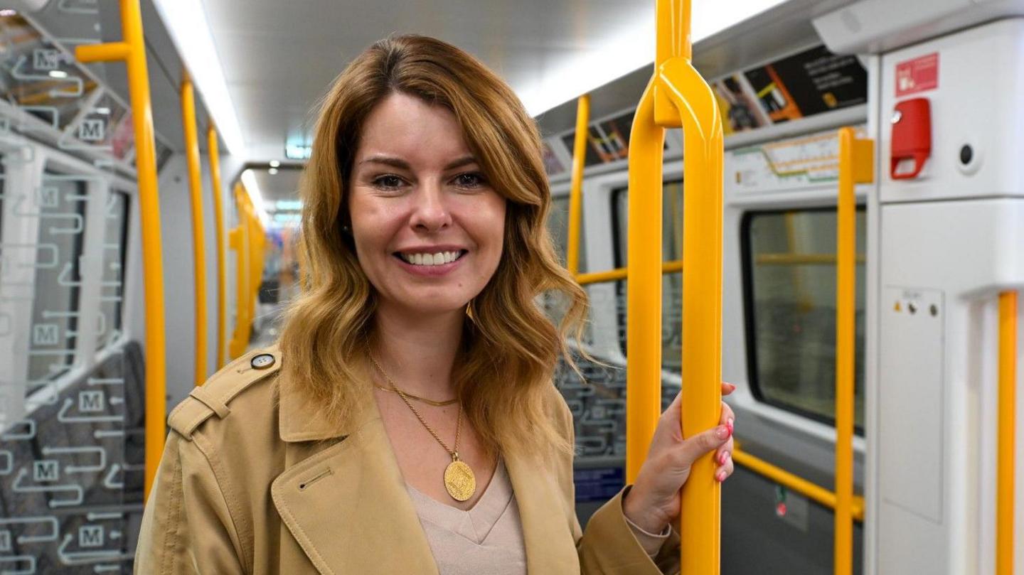 Kim McGuinness standing inside one of the new Tyne and Wear Metro trains. She has blonde hair and is wearing a beige coat. She is holding onto a yellow pole.